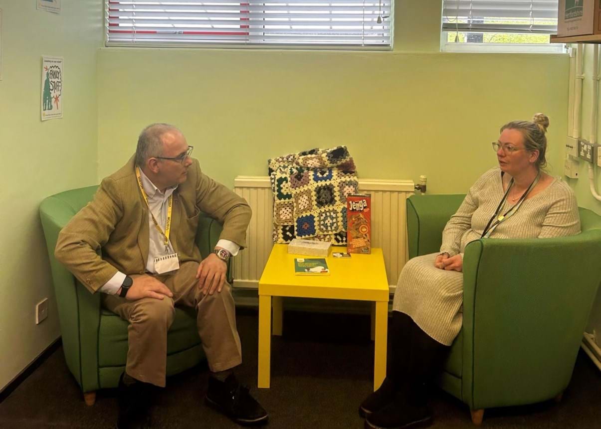 Harlow MP Rob Halfon speaking with a Place2Be School Project Manager. The pair are sitting on green armchairs inside a room with light green painted walls.