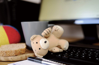 Teddy bear on a computer keyboard, next to a cup of tea and slice of toast