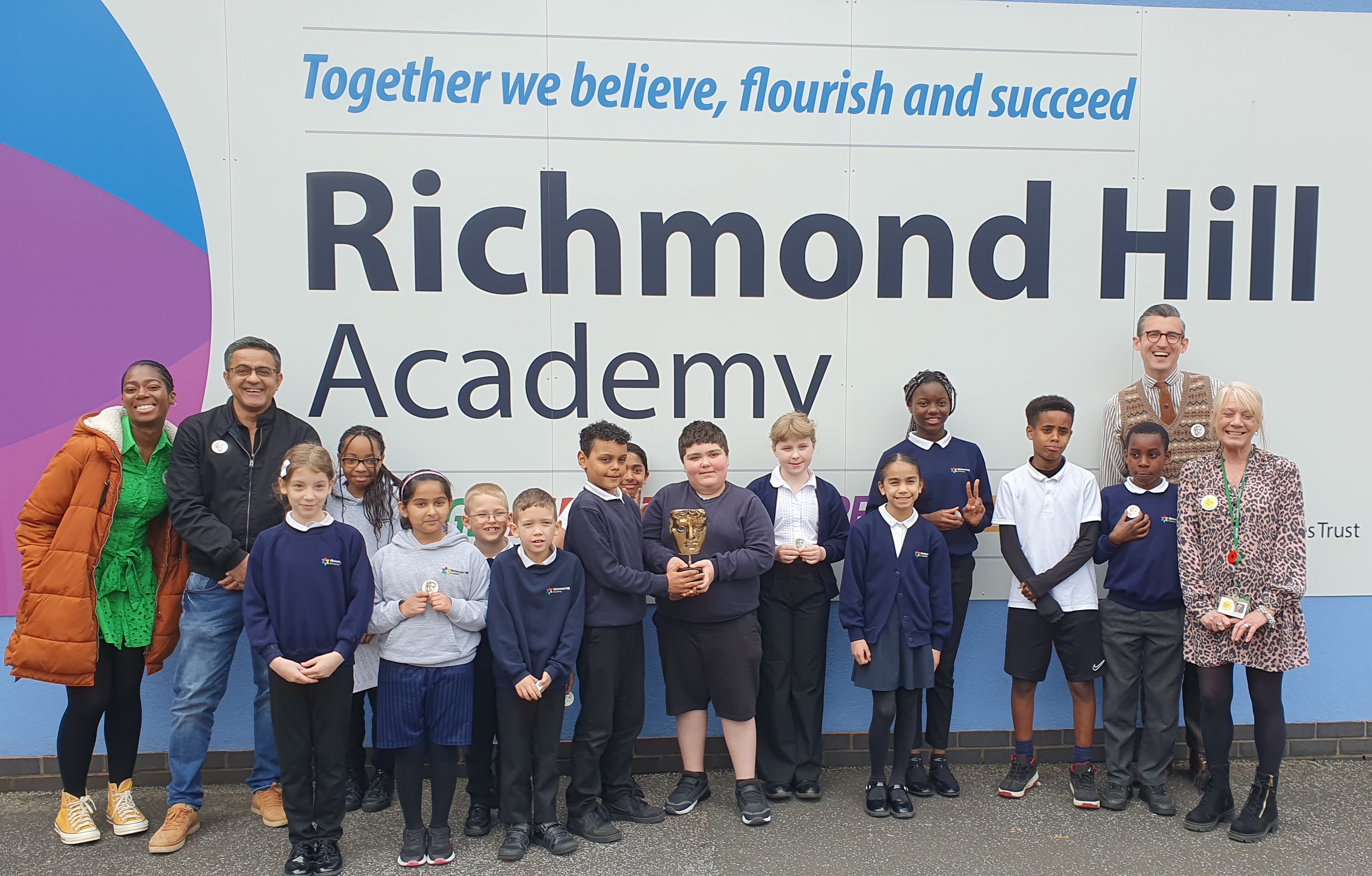 Shanequa Paris (left), Waqar Ahmed, and Ben Shires (right) standing in front of the Richmond Hill Academy sign with pupils. Two children are smiling while holding the BAFTA trophy.