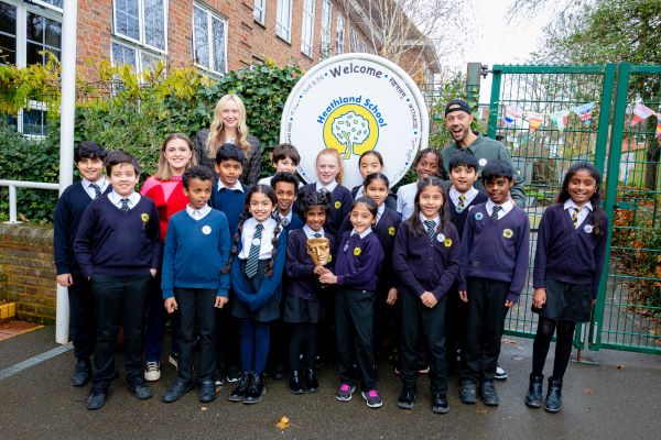 Kia Pegg (back left), Gwendoline Christie (back, second from left) and MC Grammar (back, right) standing outside with Healthland Primary School pupils. They are standing in front of the school sign, and smiling directly at the camera. Two children standing at the centre front are holding a BAFTA trophy.