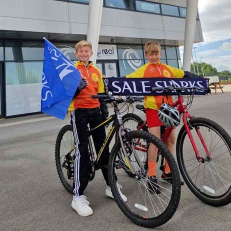 Jayden and Eric standing outside AJ Bell Stadium with a Sale Sharks flag