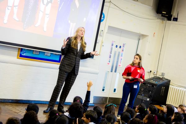 Gwendoline Christie (left) and Kia Pegg (right) presenting in assembly to the pupils at Heathland Primary School. Gwendoline is standing in the centre of hall and is talking to the children.
