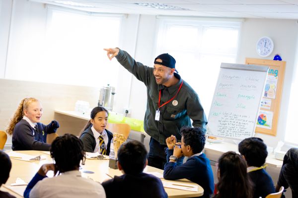 MC Grammar with pupils at Heathland Primary School during a roundtable discussion. The children are sitting in a circle and laughing. MC Grammar is standing up and making a joke to the children.