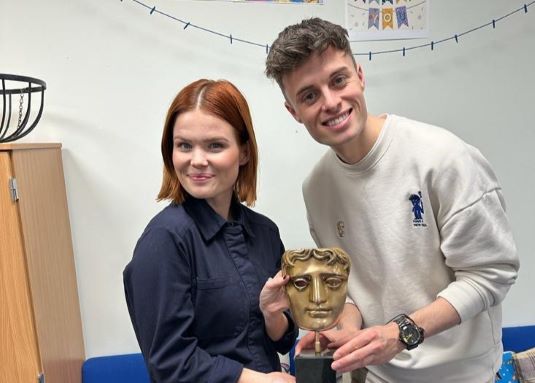 Lindsey Russell (left) and Joe Tasker (right) posing with a BAFTA Trophy. They are both looking into the camera and smiling.