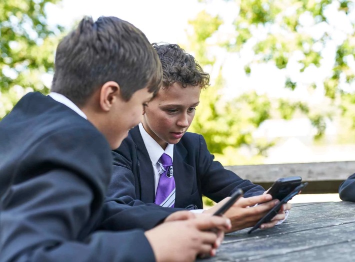 Two young boys in school uniform sit next to each other as part of a group outside. They are looking at their phones.