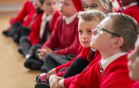 A class of primary aged pupils in red uniform sitting on the floor in a school hall