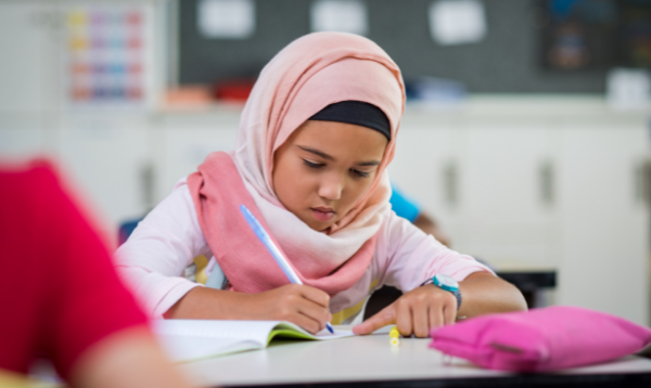 Young girl wearing hijab, writing on desk at school