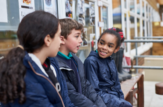 3 primary aged pupils sat in a school playground, wearing coats, smiling in conversation