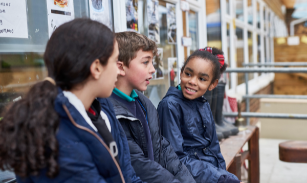 3 primary aged pupils sat in a school playground, wearing coats, smiling in conversation