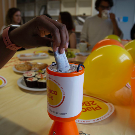 someone putting money into a Place2Be donation tin, which is sitting near Place2Be balloons on a cake sale table