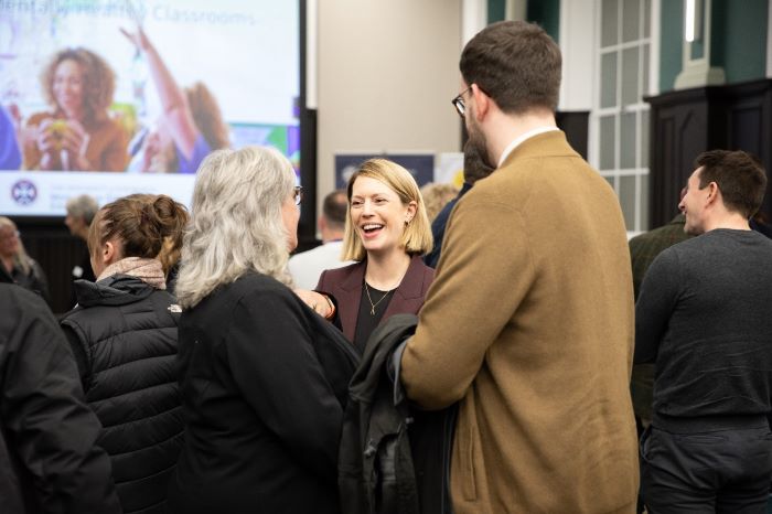 There are three people standing up and having a conversation. Two of the members are standing with their backs facing to the camera. The person in the middle is facing the camera and is smiling at the person who is standing on the left.