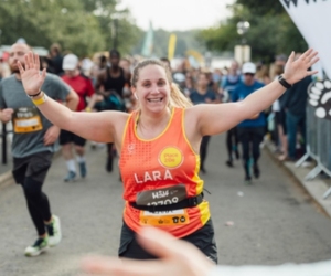 Woman in a Place2be running vest, taking part in a race and running past with her arms in the air.