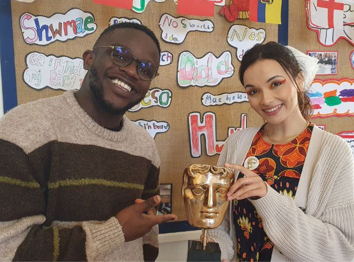 De-Graft Mensah and Mirain Iwerydd standing in a classroom smiling while holding a BAFTA trophy. De-Graft Mensah and Mirain Iwerydd standing in a classroom smiling while holding a BAFTA trophy.