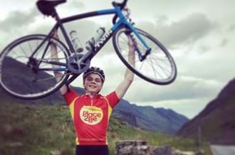 Tom wearing a Place2Be cycling top and holding his bike above his head with mountains in the background. He is smiling at the camera.