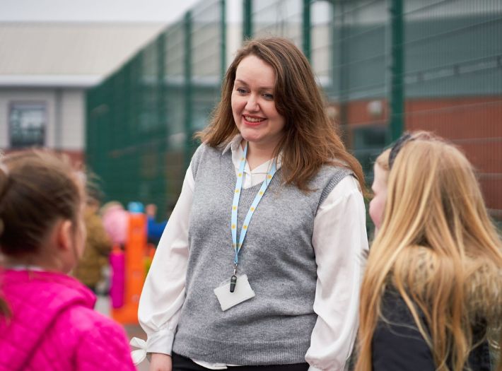 Two young students facing a woman who is smiling at them outside on school grounds. She is wearing a grey vest and a Place2Be lanyard.