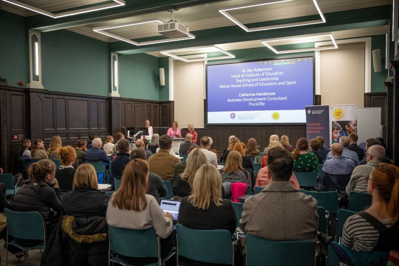 A group of people sitting in a room. They are watching and listening to a presentation delivered by Moray House and Place2Be. They have their backs to the camera.