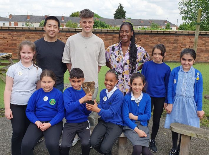 Brandon Cheung, Joe Tasker and Shanequa Paris standing outdoors with smiling children at Pear Tree Community Junior School. 
