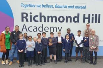 Shanequa Paris (left), Waqar Ahmed, and Ben Shires (right) standing in front of the Richmond Hill Academy sign with pupils. Two children are smiling while holding the BAFTA trophy. 