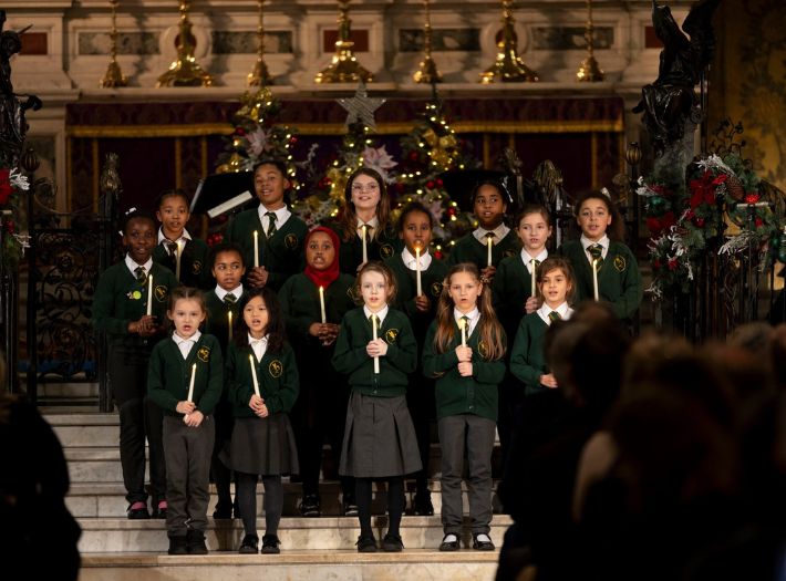 The Goldfinch Primary School choir performing at Holy Trinity Church in Sloane Square. Fifteen children dressed in school uniform are holding candles while singing.