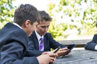 Two young boys in school uniform sit next to each other as part of a group outside. They are looking at their phones.