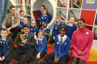 BAFTA Roadshow presenters Sam Homewood (left) and Shanequa Paris (right) sit in a classroom with a group of smiling children from Ark Castledown Primary Academy in Hastings.