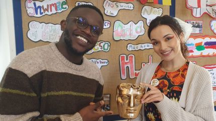  De-Graft Mensah and Mirain Iwerydd standing in a classroom smiling while holding a BAFTA trophy. 