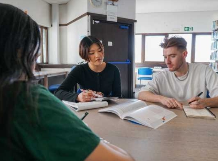 Three university students sitting together looking at a textbook and making notes. They are sitting in a library,