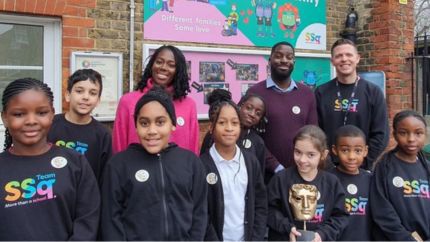 Presenters Shanequa Paris and Inel Tomlinson photographed smiling outdoors with Surrey Square Primary students and Head Teacher Matt Morden. 