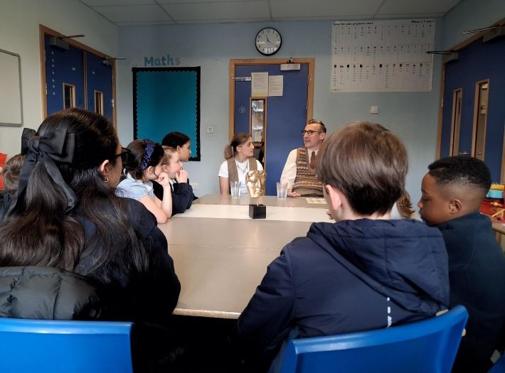 Kira Pegg and Ben Shires sitting with pupils from Sacred Heart Primary School around a table in a classroom. They are all in discussion with a BAFTA trophy in the middle of the table.