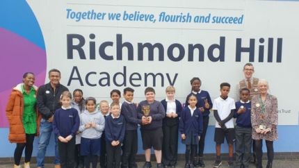 Shanequa Paris (left), Waqar Ahmed, and Ben Shires (right) standing in front of the Richmond Hill Academy sign with pupils. Two children are smiling while holding the BAFTA trophy. 