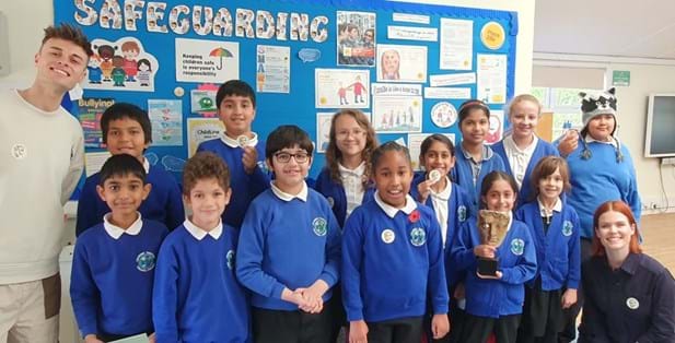 Pupils at Chalkhill Primary School with Joe Tasker (left) and Lindsey Russell (right). They are all smiling at the camera. A child standing in the front row is holding a BAFTA Trophy.