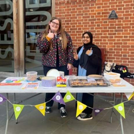 Two women standing behind a cake sale table at a university, with Place2Be bunting hanging from the table.