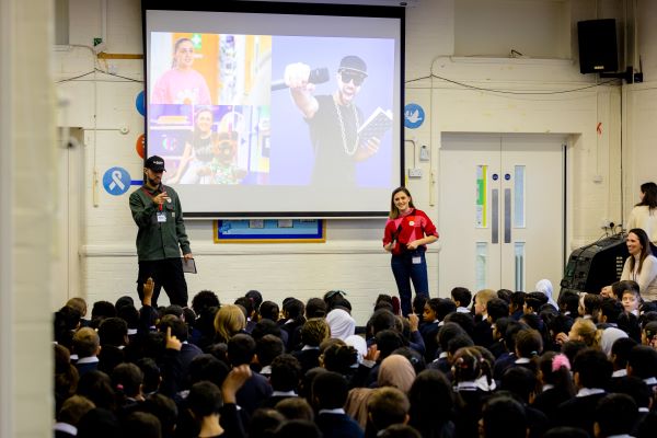 MC Grammar (left) and Kia Pegg (right) presenting the BAFTA Roadshow to pupils at Heathland Primary School. They are standing with the children facing towards them.