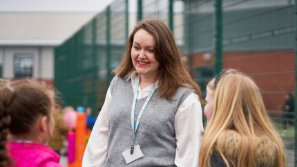 Two young students facing a woman who is smiling at them outside on school grounds. She is wearing a grey vest and a Place2Be lanyard.