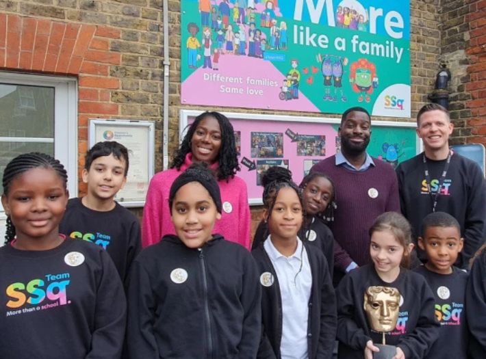 Presenters Shanequa Paris and Inel Tomlinson photographed smiling outdoors with Surrey Square Primary students and Head Teacher Matt Morden. 