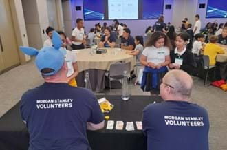 Two men wearing T-shirts saying 'Morgan Stanley Volunteers' are sitting at a table in a room with school children and teachers.