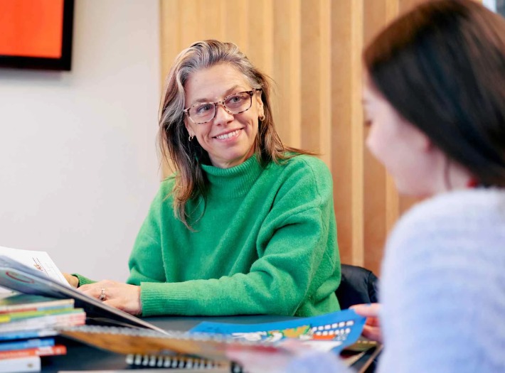 A woman in a green jumper sitting at a table with a book, talking to another woman. 
