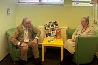 Harlow MP Rob Halfon speaking with a Place2Be School Project Manager. The pair are sitting on green armchairs inside a room with light green painted walls.