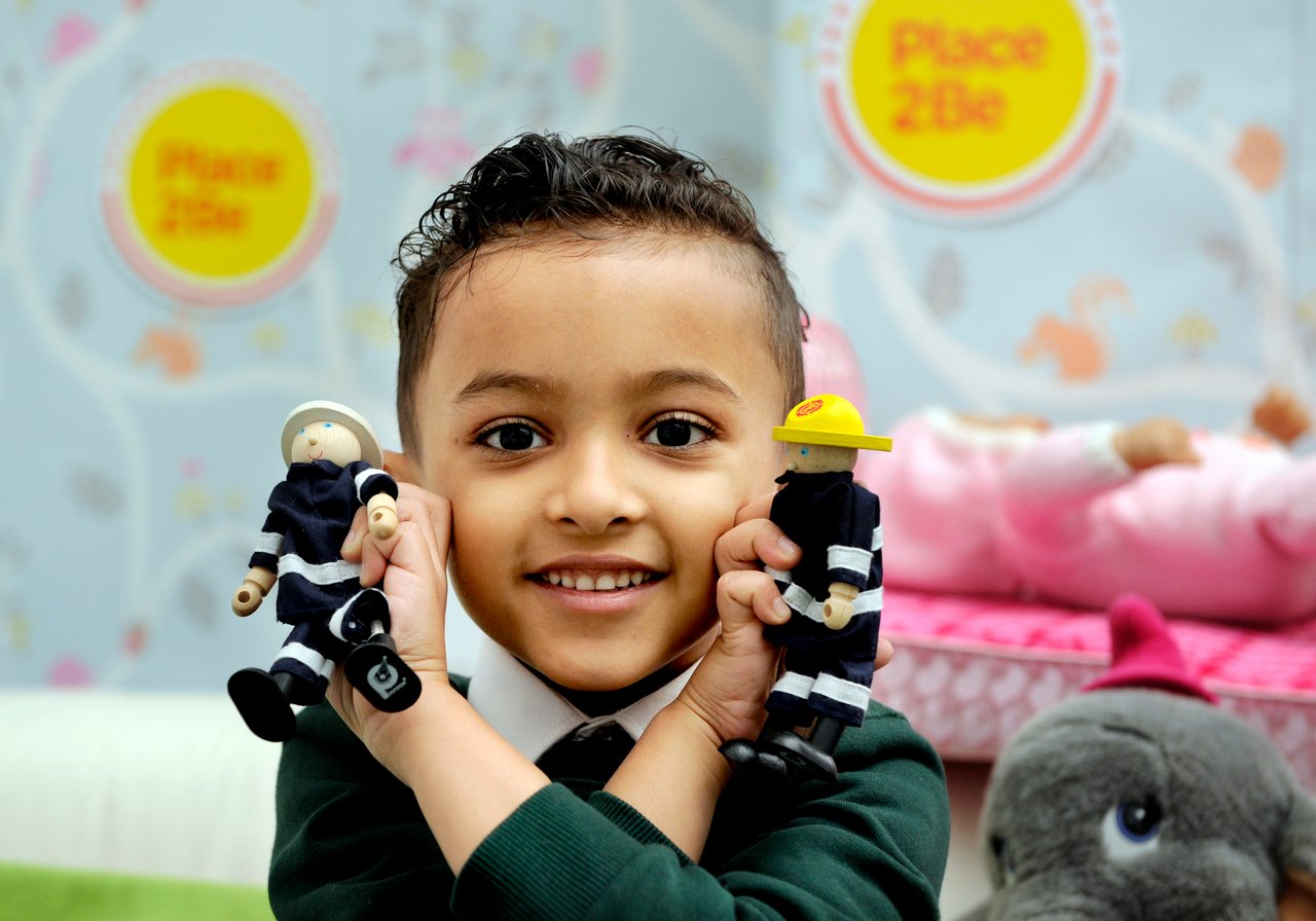 Smiling boy holds up two fireman toys with Place2Be posters in background