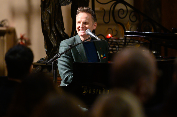 Joe Stilgoe performing at the Place2Be carol concert. He is sitting at the piano and smiling at the crowd.
