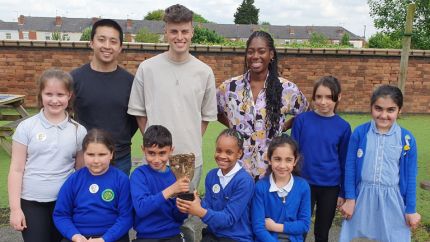Brandon Cheung, Joe Tasker and Shanequa Paris standing outdoors with smiling children at Pear Tree Community Junior School. 