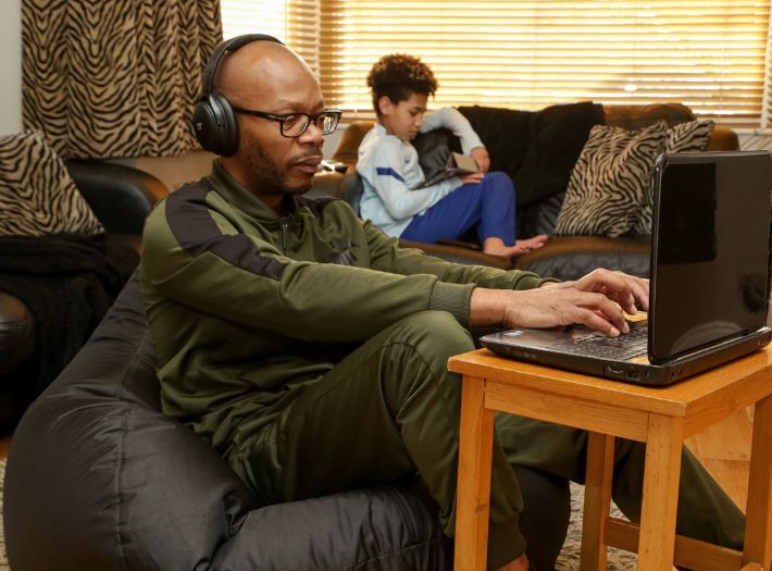 A parent and child sitting at home. The parent is wearing headphones and using a laptop and the child is sitting on the sofa.