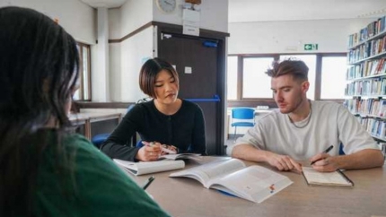 Three university students sitting together looking at a textbook and making notes. They are sitting in a library,
