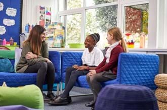 A woman sat on a sofa, smiling at two young girls wearing school uniform sat opposite her on another sofa. They are in a school classroom.